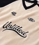 Suéter de punto con corte boxy, diseño bicolor en crema y negro. Presenta cuello en V, mangas largas con rayas laterales y estampa frontal con lettering Untitled y detalles gráficos. Cuenta con terminaciones acanaladas a rayas en el ruedo y puños.