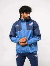 Campera de lluvia Umbro del Club Nacional de Football, con diseño de bloques de color en tonos azul y celeste. Cuenta con capucha, cierre frontal completo, puños elásticos y detalles de piping. Incluye el logo de la marca y el escudo del club en el pecho.