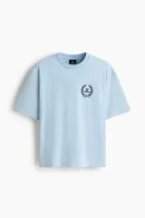 Remera de algodón color celeste, con cuello redondo, mangas cortas y corte holgado. Presenta un pequeño estampado frontal en el pecho con diseño de corona de laureles y texto.