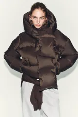 Campera puffer corta de color marrón oscuro, con diseño acolchado, cuello alto y capucha integrada. Presenta cierre frontal con botones magnéticos y mangas largas con puños elásticos.