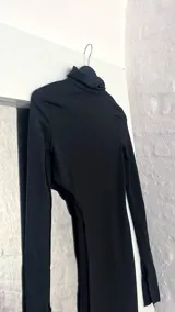 Top negro de manga larga con cuello alto y diseño asimétrico que deja al descubierto un costado del abdomen y cae en un panel largo hasta la rodilla o tobillo.