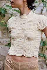 Top de punto con mezcla de viscosa, color beige, con cuello subido y manga corta. Presenta un diseño con frunces verticales y detalles bordados a tono, con terminación de ruedo ondulado.
