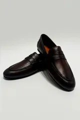 Mocasín de cuero color café, diseño clásico de estilo penny loafer con suela plana y costuras visibles.