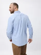 Camisa de jean manga larga en color azul claro, con cuello clásico, cierre frontal con botones y un bolsillo en el pecho con detalle de etiqueta.