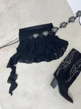 Minifalda de encaje negro con diseño de volados y una tira lateral larga y asimétrica. Presenta un cinturón incorporado con eslabones metálicos en forma de corazón.