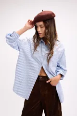 Camisa de corte oversize con estampado de rayas verticales finas en color azul sobre fondo claro. Presenta cuello clásico, cierre frontal con botones y un bolsillo aplicado en el pecho.