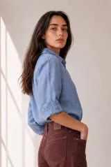 Blusa azul de lino con cuello camisero, escote en V y mangas tres cuartos con vuelta. Presenta un diseño holgado con dobladillo redondeado y un pequeño parche con logo bordado en el lateral inferior.