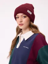 Gorro tipo beanie de punto acanalado color bordó, con etiqueta rectangular blanca con logo de la marca en el frente.