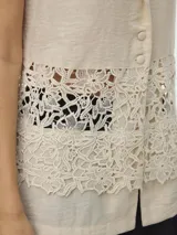 Top sin mangas color avena (oatmeal) con cuello en V y solapas tipo blazer. Presenta un panel central de encaje floral calado que deja ver la piel o la prenda interior.