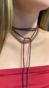 Gargantilla tipo choker que combina una hilera de cuentas metálicas plateadas con un lazo largo de gamuza en color bordó, diseñado para anudarse al cuello.