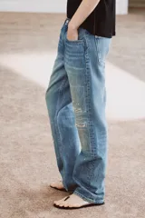 Jeans de tiro medio con corte recto y largo completo. Presentan un diseño de cinco bolsillos, cierre frontal con cremallera y botón, y un acabado con efecto lavado. Incluyen detalles de roturas y parches decorativos en la parte delantera.