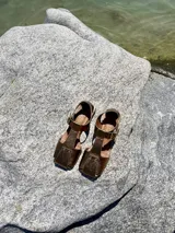 Sandalias de cuero color bronce, con diseño de punta cuadrada, taco cuadrado símil madera y tira ajustable con hebilla en el tobillo.