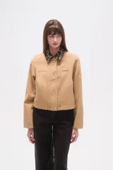 Campera corta color beige, con cuello camisero estampado floral en tonos oscuros y cierre frontal de botones a presión. Presenta bolsillos superiores con solapa y pespuntes verticales en el frente.
