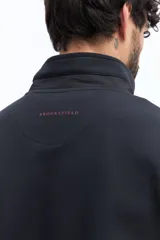 Campera negra de algodón con cierre frontal completo, cuello alto y bolsillos laterales con botón a presión. Presenta un pequeño parche rectangular con texto en el pecho izquierdo.