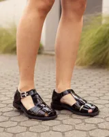 Sandalias de cuero negro charolado estilo romano, con tiras entrelazadas, tira en T y hebilla metálica de ajuste lateral.