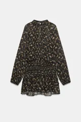 Vestido corto de cuello subido y manga larga con puños elásticos. Presenta cintura elástica con detalle de nido de abeja, abertura frontal y trasera, y bajo con volantes. Cierre posterior con botones.