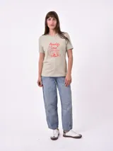 Remera de manga corta color beige claro con cuello redondo. Presenta un estampado gráfico central con un plato de pasta rodeado por un borde azul y blanco, sobre un fondo de franjas verticales rojas y blancas. El estampado incluye texto en cursiva en tonos turquesa y rojo.