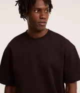 Remera masculina de corte boxy, color marrón oscuro, con cuello redondo y mangas cortas. Confeccionada en malla pesada con textura gomosa.