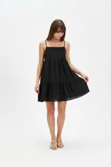 Vestido corto negro de algodón con diseño holgado, escote cuadrado y tirantes finos. Presenta detalles de bordado con perforaciones en toda la prenda y falda con volados en capas.