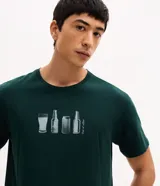 Remera de algodón color verde oscuro con cuello redondo y manga corta. Presenta una estampa frontal pequeña y centrada con ilustraciones de vasos y botellas de cerveza junto a la frase 'Out of office'.