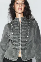 Chaqueta entallada de terciopelo color gris con efecto acid wash, cuello subido, manga larga y cierre frontal con ganchos metálicos. Presenta detalles de pasamanería con falsos botones en el delantero de inspiración militar.
