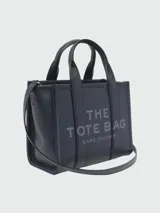 Bolso tote pequeño de cuero texturizado color gris con doble asa corta superior y correa larga extraíble y ajustable para llevar al hombro. Estampado frontal con el texto "The Tote Bag" y el nombre de la marca.