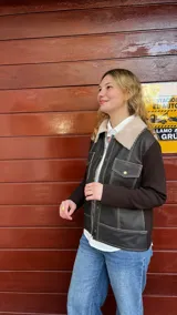Campera de diseño híbrido con cuerpo de cuero marrón y mangas de lana merino, cuello con solapas de corderito sintético, cierre frontal con botones a presión y bolsillos frontales con solapa.