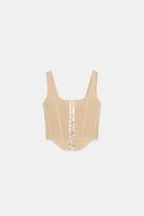 Top corsetero de tul semitransparente color beige, con escote cuadrado, tirantes anchos y cierre frontal con ganchos metálicos. Presenta bajo asimétrico y costuras reforzadas con ribetes a tono.
