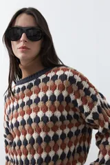 Sweater de punto con diseño geométrico de rombos en tonos azul, marrón, beige y blanco. Presenta cuello redondo, puños y ruedo acanalados en color azul oscuro, con un calce relajado y mangas largas.