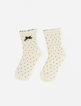 Par de medias de caña media color crema con estampado de pequeños lunares negros. Presentan un borde superior con terminación ondulada y un detalle de moña negra aplicada en uno de los laterales.