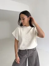 Chaleco tejido de punto color crudo, con cuello redondo y diseño de flor en relieve en el frente. Presenta un corte holgado y mangas cortas con terminación acanalada.