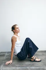 Sandalia plana negra con tira para el dedo y tira tobillera ajustable con hebilla dorada. Presenta un adorno grande de pieza metálica dorada con textura orgánica sobre el empeine.