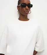Remera de algodón corte oversized, color blanco, con cuello redondo, mangas cortas amplias y ruedo asimétrico con detalle de encaje aplicado.