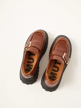 Mocasines de cuero color marrón con plataforma dentada negra. Presentan una tira frontal con hebilla metálica dorada y costuras decorativas en contraste.