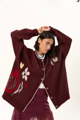 Saco de punto color vino con diseño holgado, cuello redondo y mangas largas. Presenta un gran bordado central en la espalda con forma de flor en tonos crema y rojo, acompañado de un pequeño bordado de sol y una frase bordada en la parte inferior.