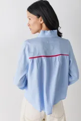 Camisa blanca de lino con cuello alto tipo gorguera, cierre frontal con botones de madera y detalles de ribetes en color mostaza en hombros y puños. Presenta volados en la botonera, cuello y puños.