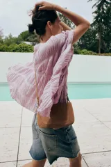 Top blanco de cuello redondo y mangas tipo capa, con textura fruncida, bajo asimétrico, lazos decorativos en el escote y en los laterales, y cierre posterior con botón.