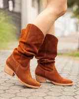 Bota estilo texana de caña alta, confeccionada en cuero gamuzado color marrón óxido. Presenta un diseño de corte en V en la parte superior de la caña, costuras decorativas y un taco bajo de madera.