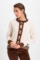 Cardigan de punto color crudo con detalles en marrón oscuro. Presenta cuello redondo, cierre frontal con botones tipo alamares y puños y ruedo acanalados con rayas horizontales. Incluye un pequeño aplique bordado en la manga.