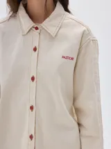 Camisa oversized de jean color crema, con botones rojos en forma de fresa y pespuntes a contratono. Tiene el logo "PASTICHE" bordado en rojo en el pecho.