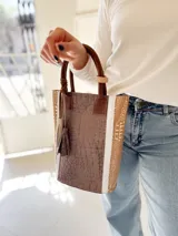 Bolso matero de cuero color taupe con textura croco y detalles en cuero color marrón. Tiene doble asa de mano y un dije de borla.