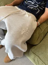 Pantalón de jogging gris melange de corte baggy, confeccionado en algodón. Presenta cintura elástica con cordón largo ajustable, bordado decorativo en la pierna izquierda y botones metálicos en la parte inferior de la botamanga para ajustar el ancho.