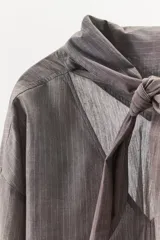 Camisa de algodón con diseño a rayas verticales, escote en pico con detalle de pañuelo ajustable mediante lazada, manga larga con puños abotonados y aberturas laterales en el bajo.
