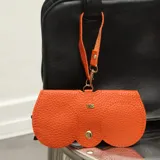 Estuche para lentes de ecocuero negro con textura granulada, diseño minimalista con cierre de broches metálicos dorados y correa de mano desmontable.