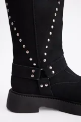 Bota de caña alta confeccionada en cuero vacuno, color negro, con diseño de tachas metálicas dispuestas en forma de arco a lo largo de la caña y correa decorativa con hebilla en la zona del tobillo. Presenta suela track de goma robusta.