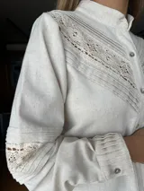 Camisa de lino color beige melange, con cuello mao, cierre frontal de botones y mangas largas. Presenta detalles de alforzas y aplicaciones de puntilla de algodón en el frente y en las mangas, con un diseño de silueta relajada.
