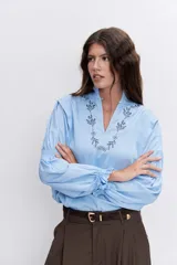 Blusa celeste de manga larga con puños fruncidos y elásticos, escote en V con cuello alto con volados y delicados bordados florales en el pecho.