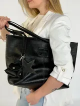 Cartera tote de cuero genuino color negro, con costuras visibles a contratono, asas dobles para hombro, parche frontal con logo grabado y charms colgantes en forma de estrella y mini monedero.
