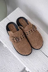 Mules de gamuza color tostado con suela negra, detalle de tira con hebilla metálica y ojales decorativos en el empeine.