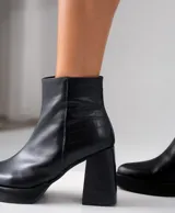 Bota corta de cuero negro con diseño combinado, presentando una textura lisa en la parte frontal y una textura tipo reptil en el talón. Cuenta con taco cuadrado alto, plataforma delantera y cierre lateral interno.